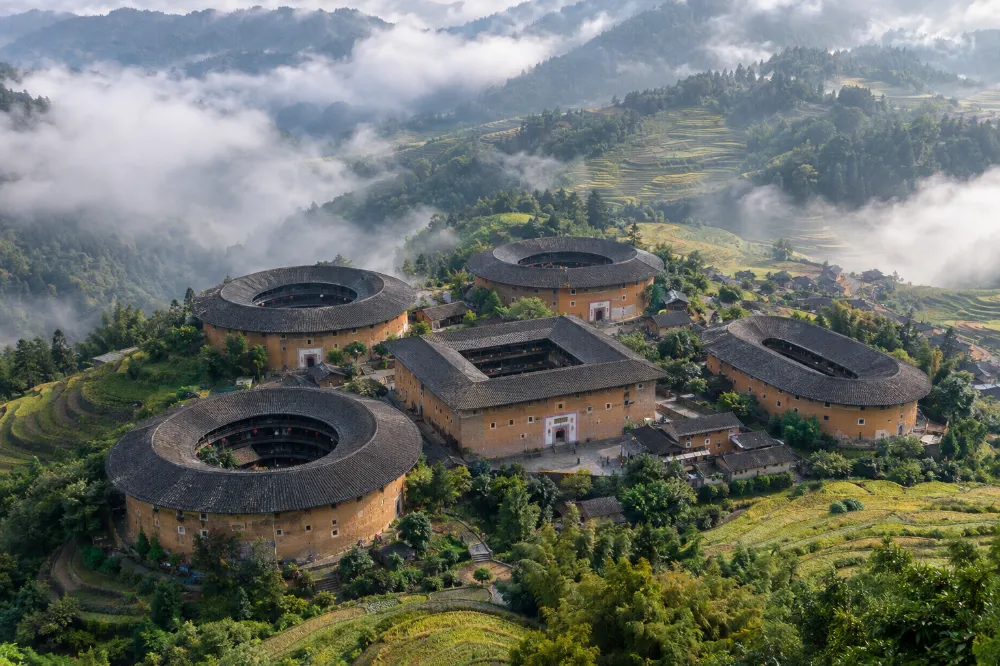 福建南靖田螺坑土樓群航拍全景,五座土樓依山勢錯落,方形步雲樓居中,三座圓樓與一座橢圓樓環繞,周圍是梯田與雲霧。 福建南靖田螺坑土樓群航拍全景,五座土樓依山勢錯落,方形步雲樓居中,三座圓樓與一座橢圓樓環繞,周圍是梯田與雲霧。